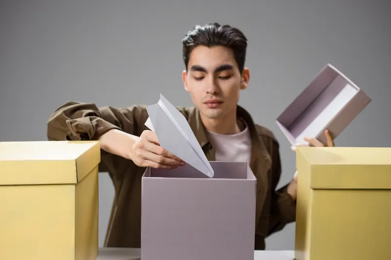 Asian man with dark hair holding a paper airplane above a large gift box indoors