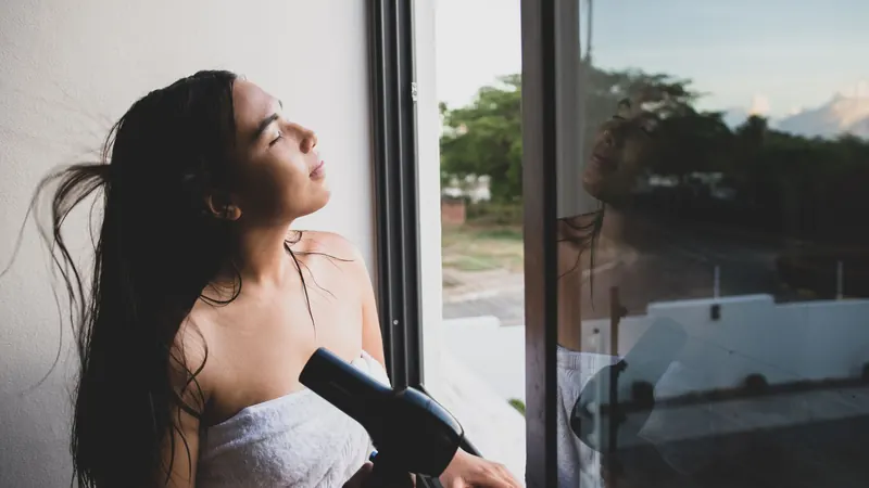 Young woman drying hair with blow dryer by open window at home