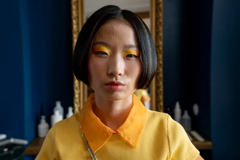 Close-up portrait of a woman with bold, artistic makeup and short black hair, wearing a bright yellow collared shirt in a hair salon