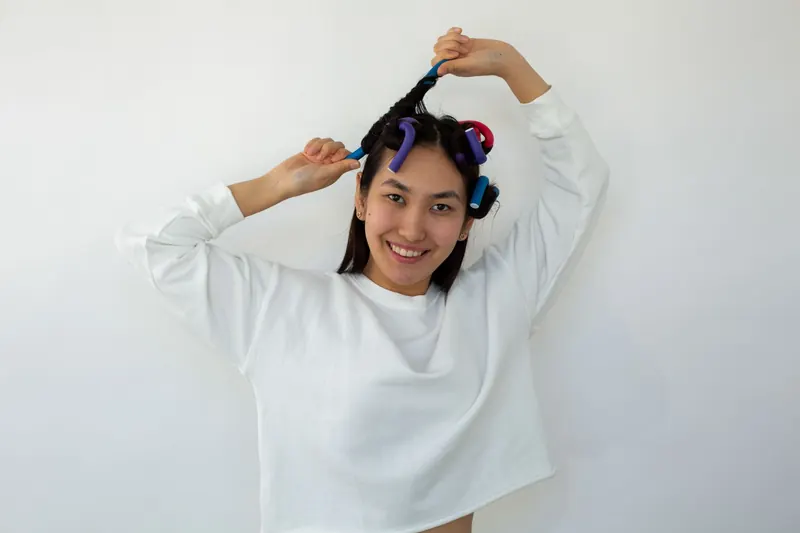 Cheerful young ethnic female looking at camera while curling black hair on roller on white background