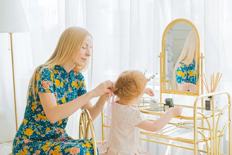 A woman braids her daughter's hair at a vanity mirror in a bright room