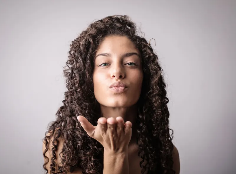 Elegant young woman with curly hair sending a playful kiss in studio. Perfect for beauty and lifestyle themes