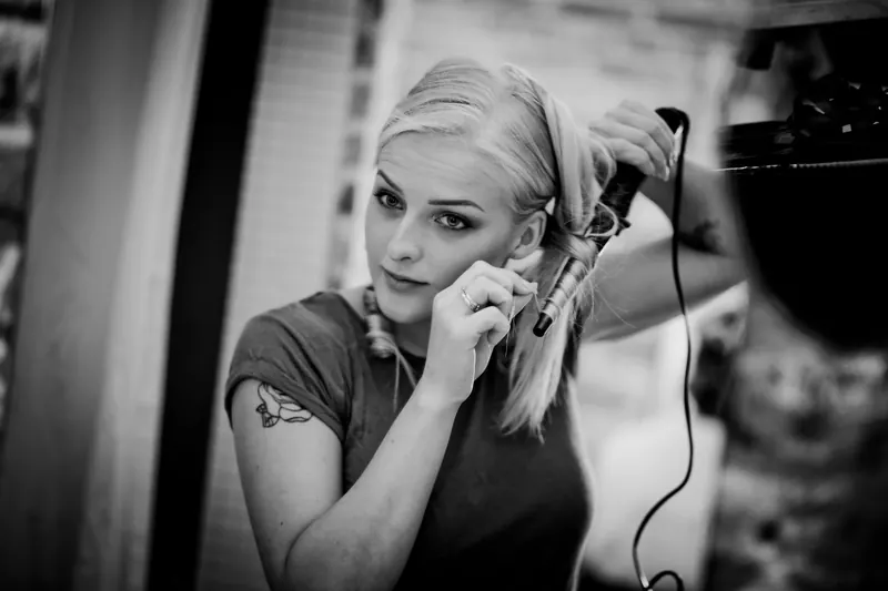 A young woman with tattoos using a curling iron to style her hair in a modern indoor setting